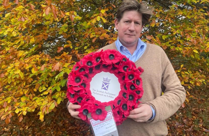 Alex stands in front of autumn foliage holding a wreath of poppies. In the centre of the wreath is the symbol of the Scottish Parliament.