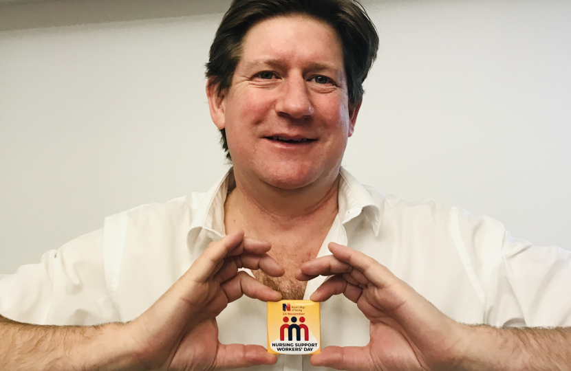 Alex poses, smiling, while holding in front of him a yellow badge that indicates Nursing Support Workers' Day.