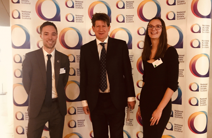 Alex with two representatives from NASS standing in front of a photo op wall with the company's logo patterned on it.