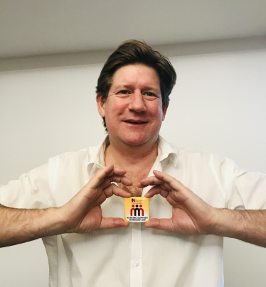 Alex poses, smiling, while holding in front of him a yellow badge that indicates Nursing Support Workers' Day.