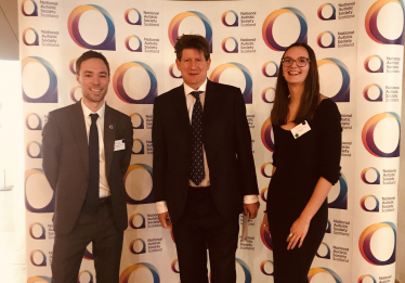 Alex with two representatives from NASS standing in front of a photo op wall with the company's logo patterned on it.